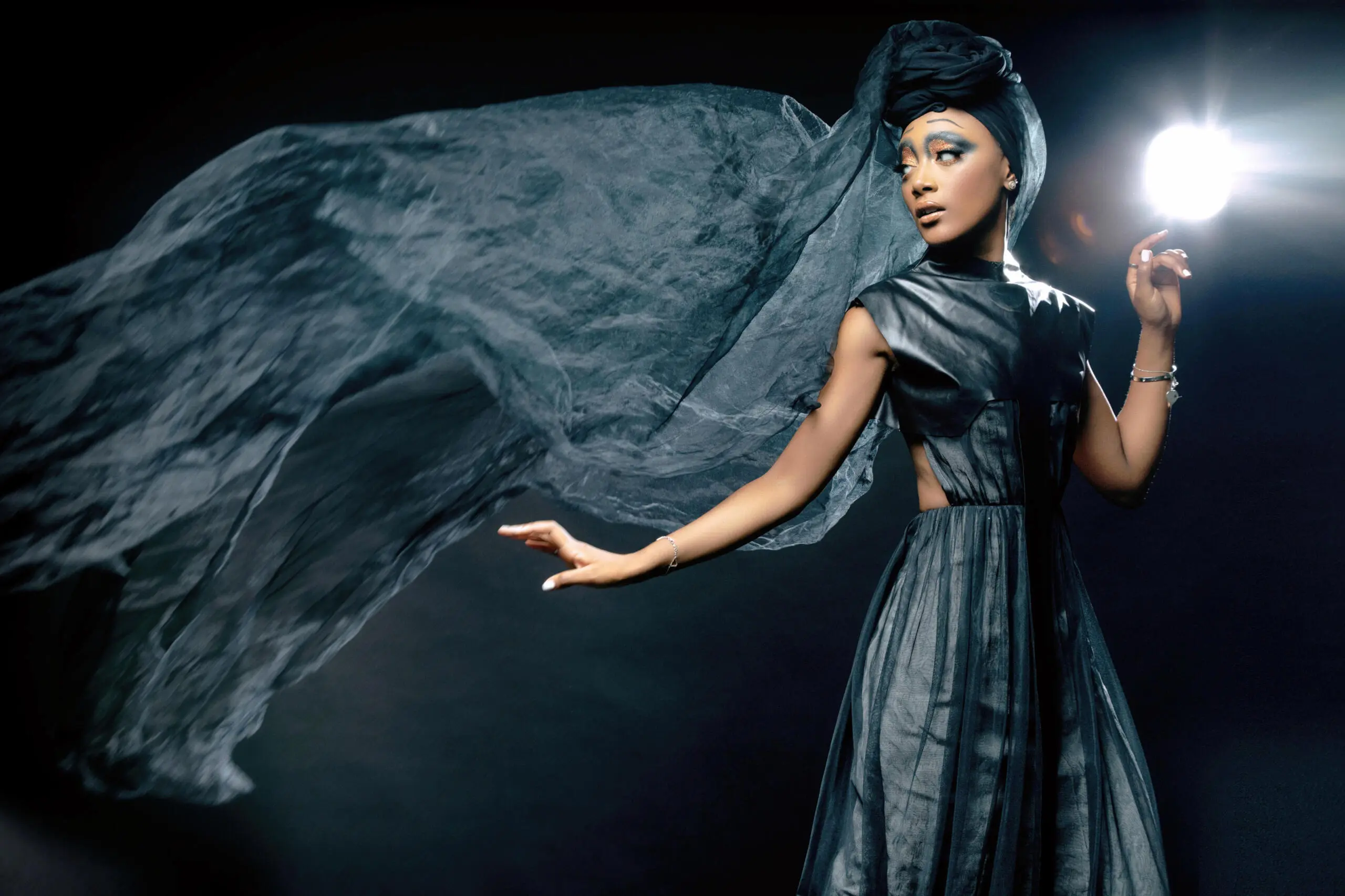 Model in a draped grey gown releasing a sweep of sheer fabric under a single studio light