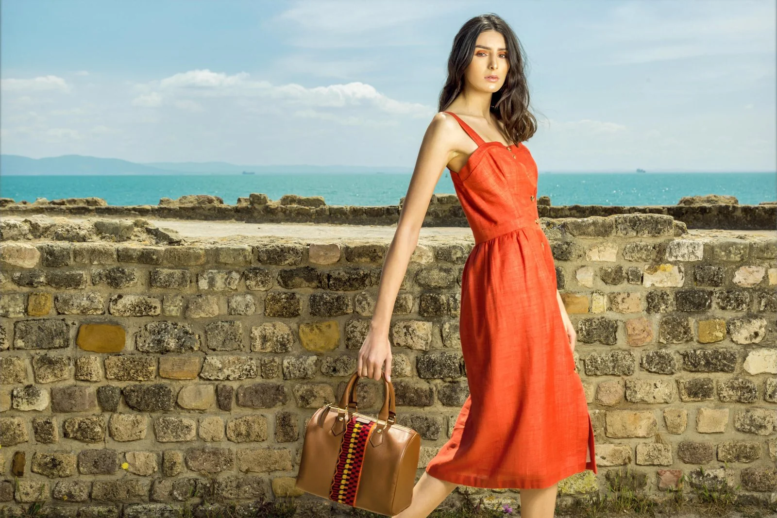 Model in an orange summer dress holding a leather bag beside a stone wall with the sea behind