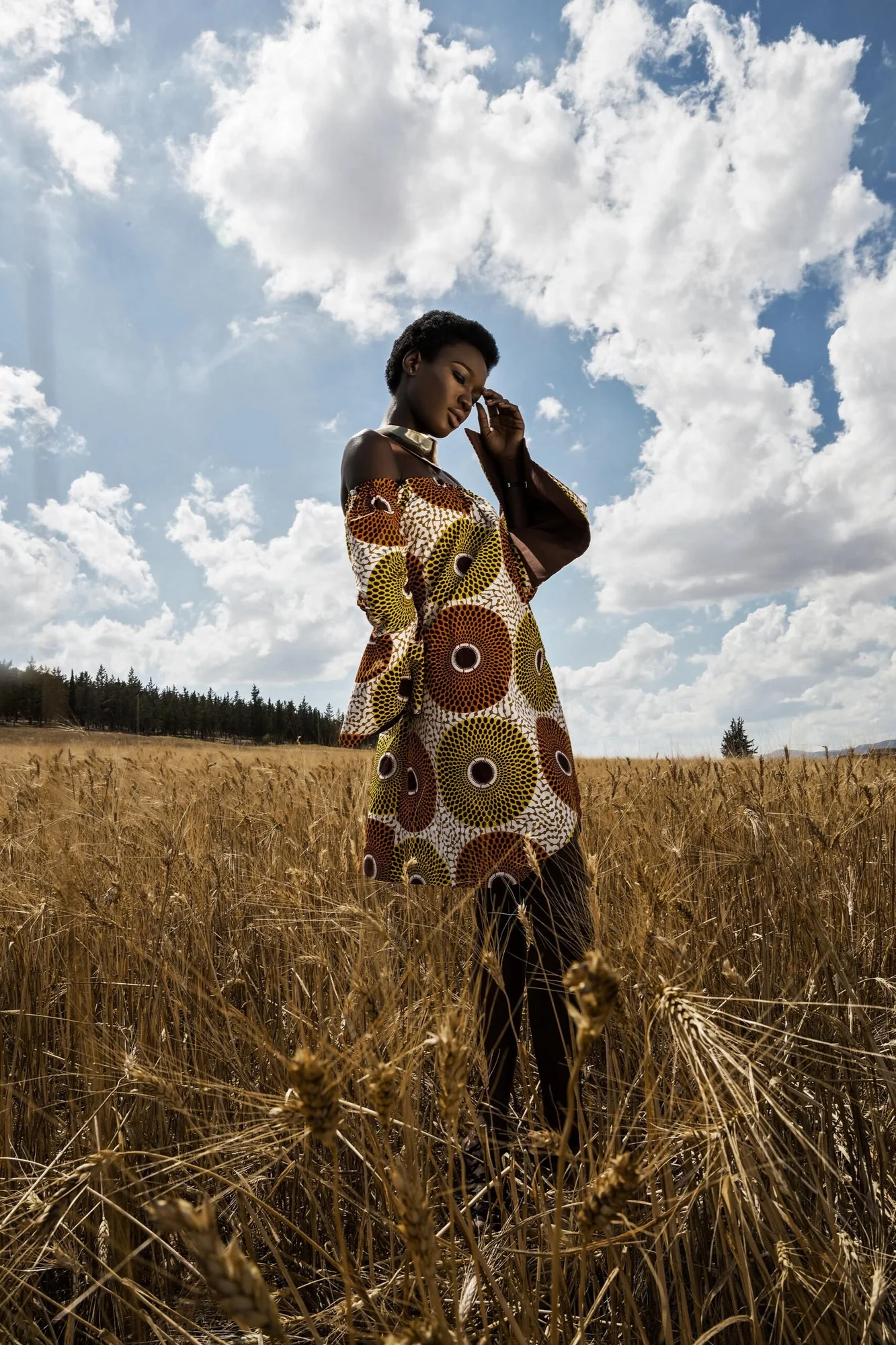 Model in a patterned coat standing in a golden field under a cloudy sky