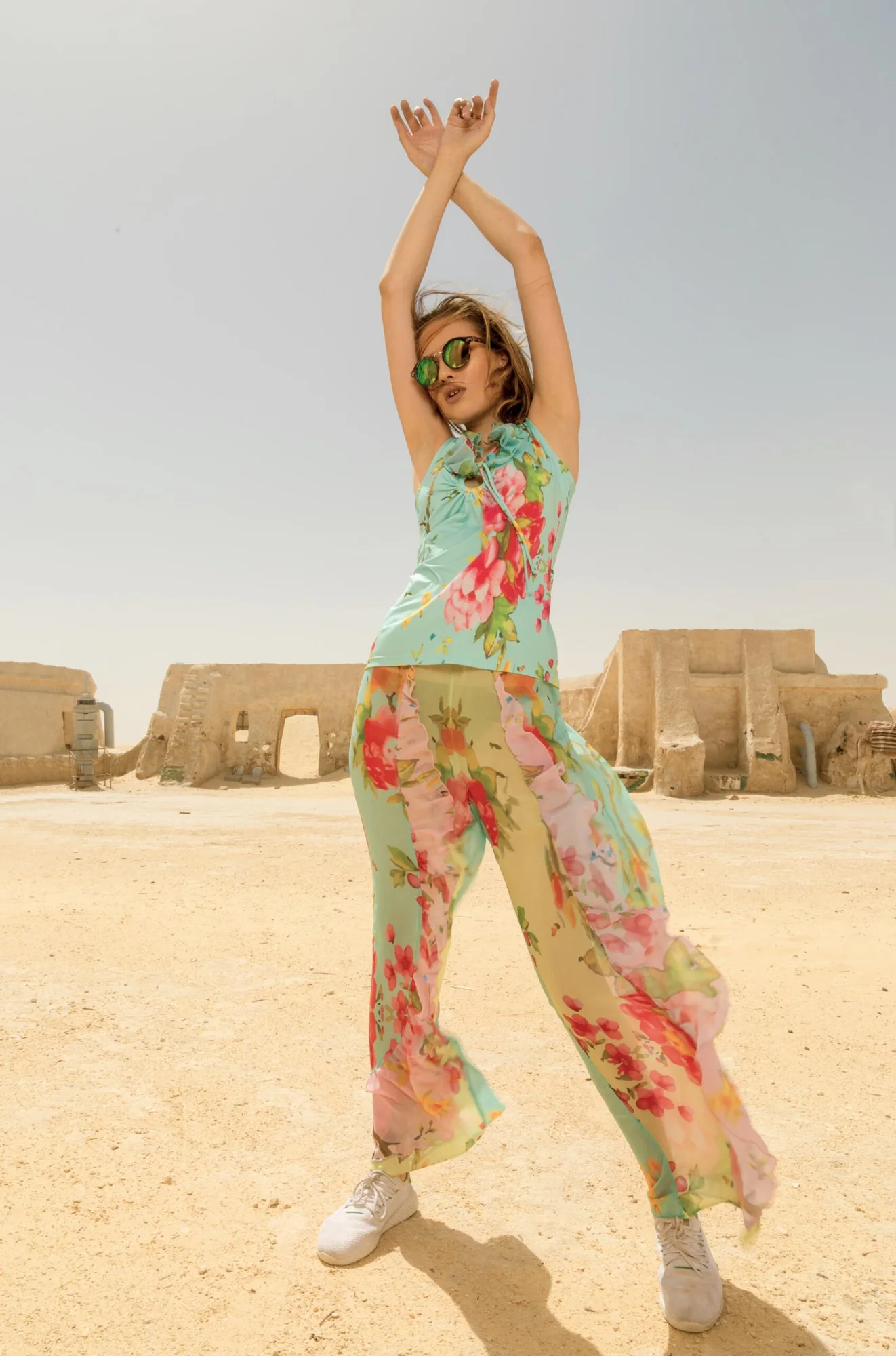 Model in a printed jumpsuit in warm sunlight on sand, with low earth-coloured buildings behind