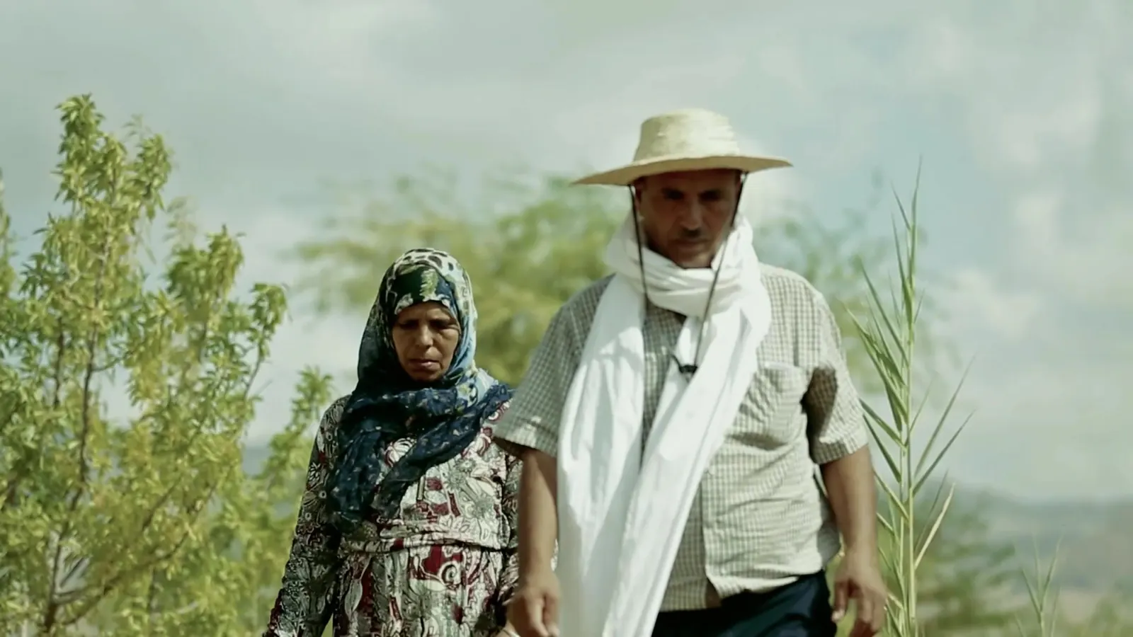 A woman and a man in a straw hat walking through green farmland, opening frame of the ILO series teaser