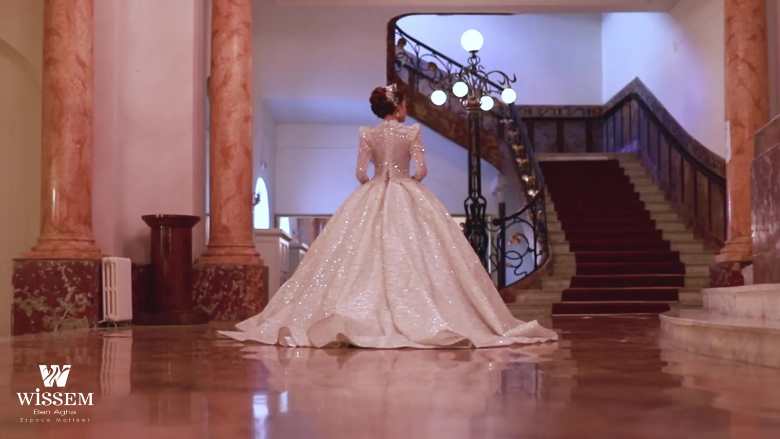 Bride seen from behind at the foot of a grand staircase in a marble hall, long gown spread on the floor