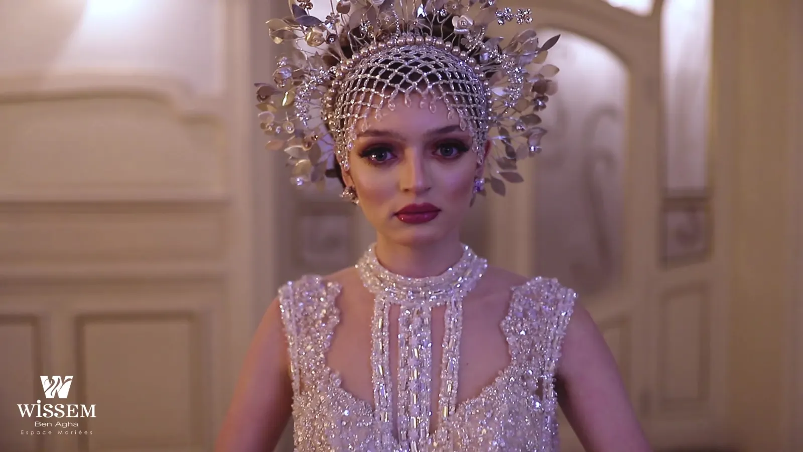 Model wearing an ornate silver crown headpiece and beaded veil, looking down past the camera