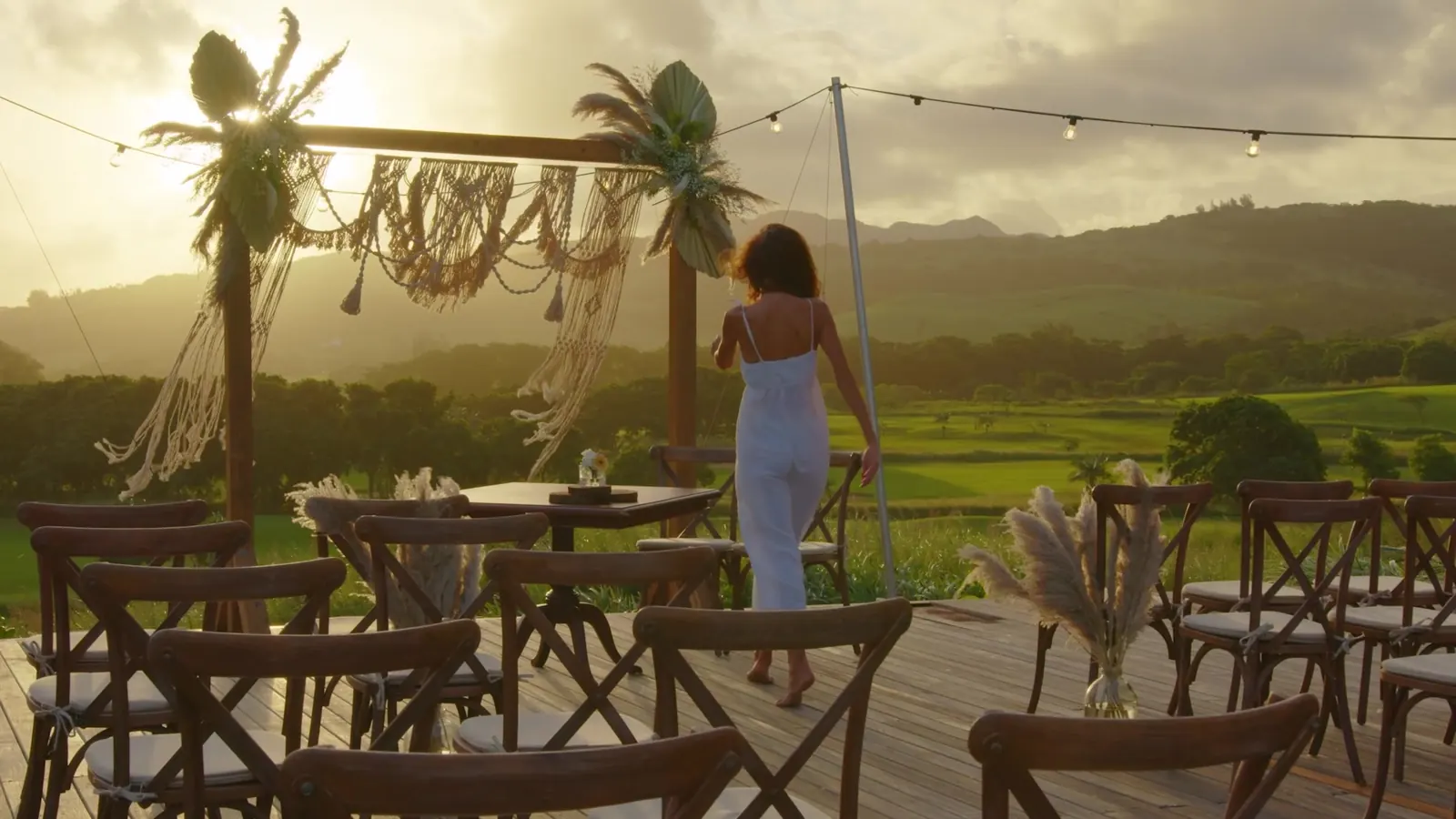 Woman in a blue dress walking under a string of lights at a hillside dining terrace at golden hour, still from the Maya Paris campaign film