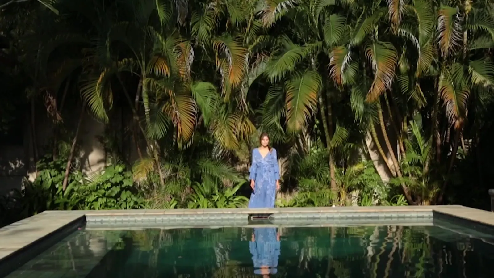 Woman in a blue dress standing at the edge of a pool framed by palms, still from the Maya Paris campaign film